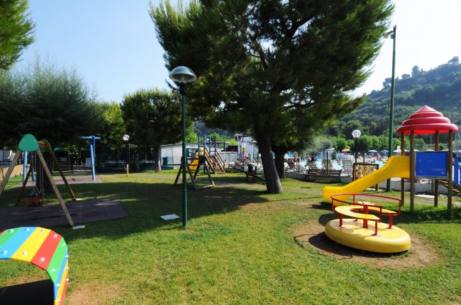 Parco giochi con scivolo giallo e altalene, circondato da alberi.