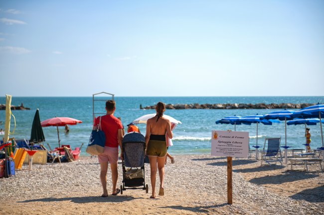 Famiglia con passeggino cammina verso una spiaggia con ombrelloni colorati e mare sullo sfondo.