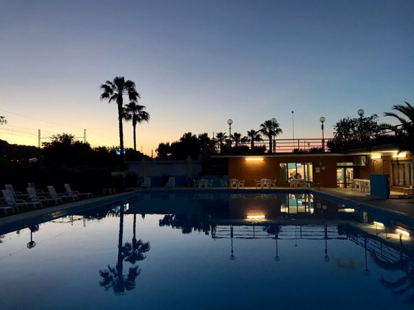 Piscina al tramonto con luci accese e palme riflesse nell'acqua.