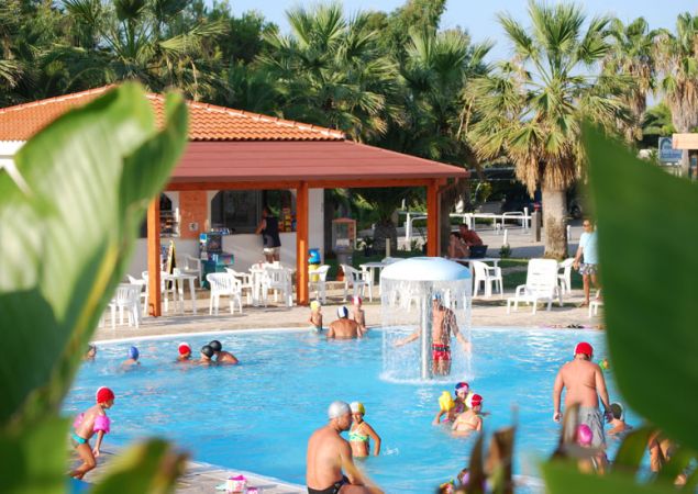 Persone che nuotano sotto una fontana in piscina, con palme e una struttura con tetto rosso.