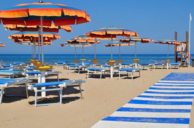 Spiaggia vuota con lettini blu e ombrelloni arancioni, passerella bianca e blu verso il mare.