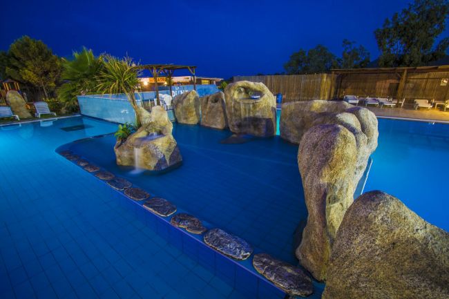 Piscina illuminata di sera con grandi rocce decorative e piante.