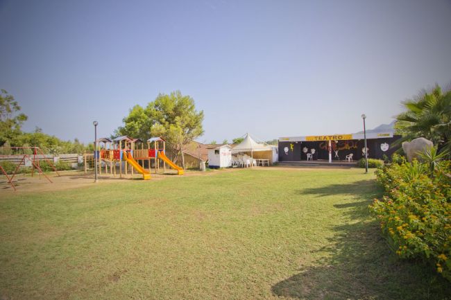 Un parco giochi con scivoli e altalene su un prato, vicino a un teatro all'aperto.