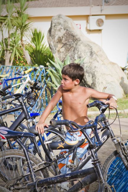 Bambino in costume da bagno vicino a biciclette parcheggiate. Sfondo con piante e rocce.