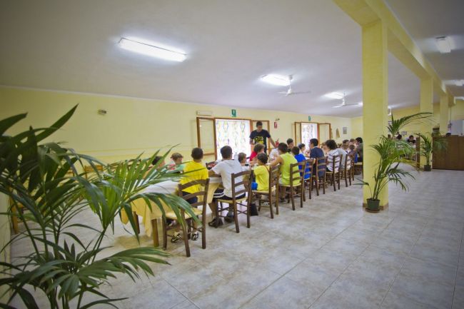 Una sala con bambini seduti a tavola. Pareti gialle e pavimento piastrellato.