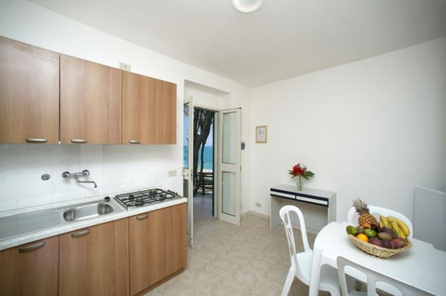 Cucina con mobili in legno, tavolo bianco con cestino di frutta, porta aperta vista mare.