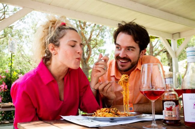 Due persone sorridenti sedute a un tavolo all'aperto, mangiano spaghetti e bevono vino.