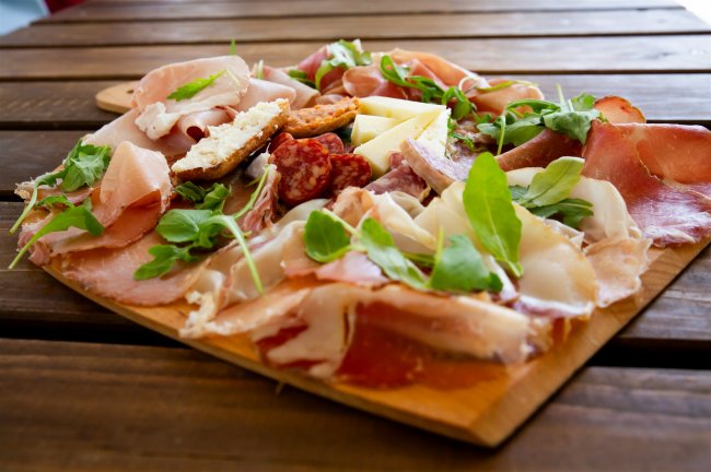 Un tagliere di legno con affettati, formaggi e rucola su un tavolo di legno.