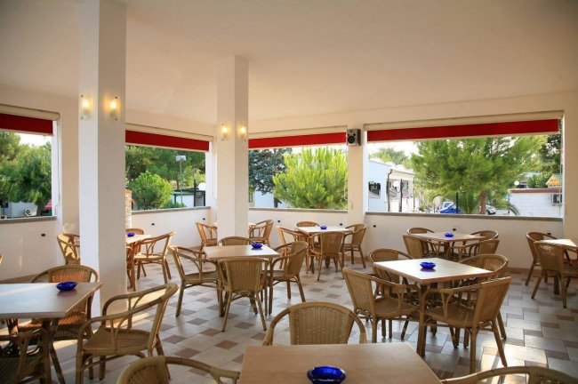 Caffetteria all'aperto con sedie e tavoli di vimini. Grandi finestre aperte con vista sugli alberi.