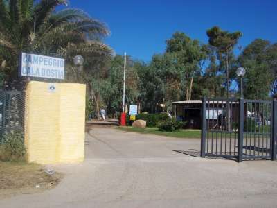 Ingresso di un campeggio con cancello aperto e cartello, circondato da alberi e palme.