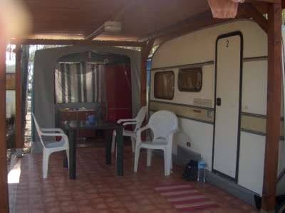 Caravan e area esterna coperta con sedie bianche e tavolo su pavimento piastrellato.