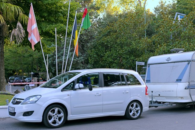 Auto bianca che traina una roulotte, vicino a bandiere e alberi verdi.