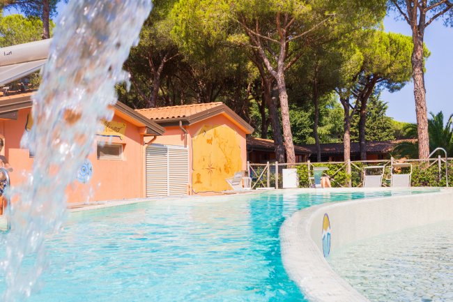Piscina con cascata d'acqua, circondata da alberi e un edificio arancione sullo sfondo.