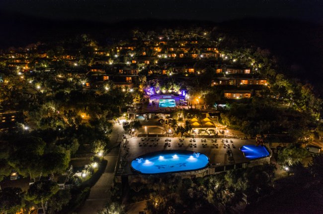 Vista notturna di un resort con piscine illuminate e capanne sparse su una collina.