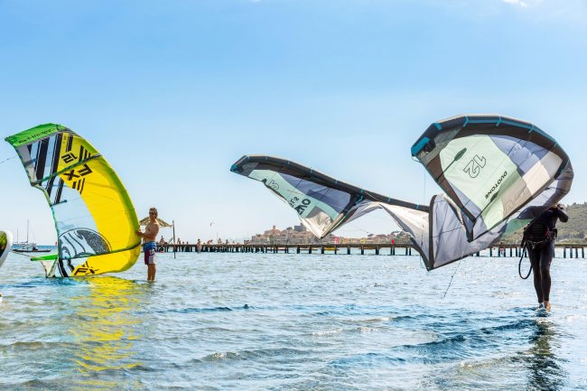Persone fanno kitesurf in mare, con un molo sullo sfondo.