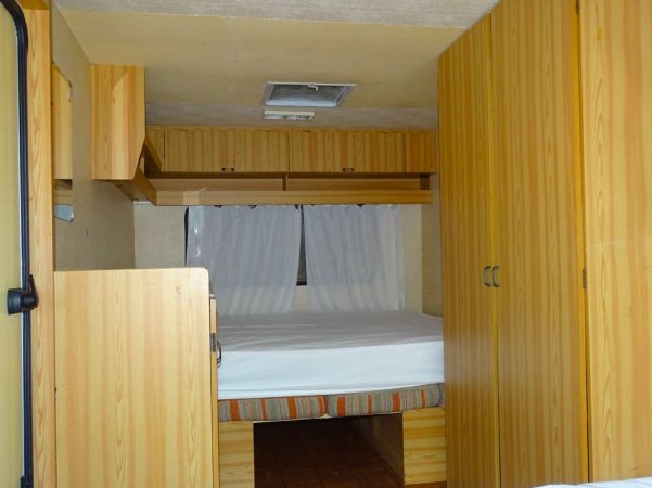 Interno di una roulotte con letto matrimoniale e armadi in legno chiaro.