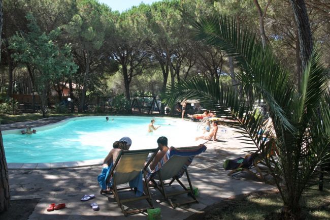Persone su sedie a sdraio e bambini in una piscina circondata da alberi.