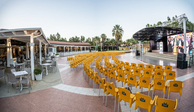 Sedie gialle disposte davanti a un palco all'aperto con tavolini.