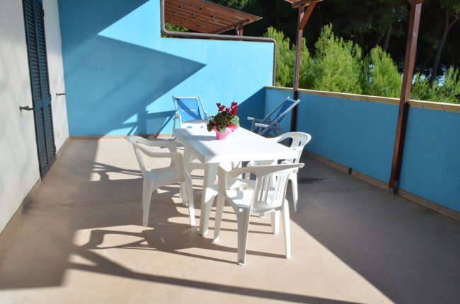 Ampia terrazza con tavolo e sedie bianche, pianta su tavolo, diverse sedie a sdraio blu.