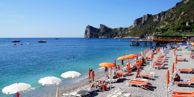 Spiaggia con lettini, ombrelloni arancioni e bianchi, vista mare e scogliera.