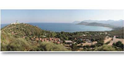 Ampio panorama costiero con colline verdi e villaggio. Mare e montagne sullo sfondo.