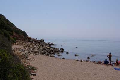 Spiaggia con sassi e mare calmo, alcune persone sulla battigia, vegetazione a sinistra.