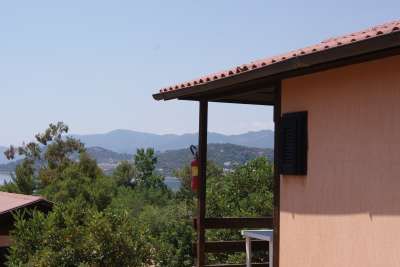 Un edificio beige con un piccolo balcone, circondato da vegetazione e montagne distanti.