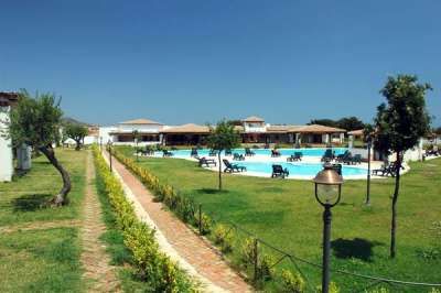 Un prato verde conduce a una piscina con sdraio. Edifici bassi in lontananza sotto un cielo blu.
