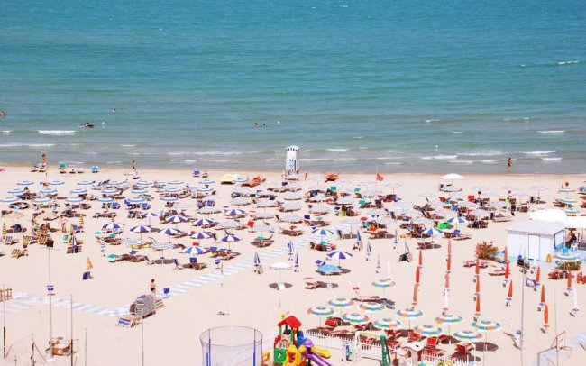 Vista aerea di una spiaggia affollata con ombrelloni colorati e mare in lontananza.
