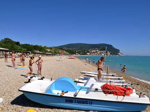 Spiaggia con pedalò, persone in costume e colline verdi sullo sfondo.