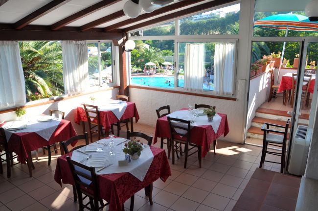 Una sala da pranzo luminosa con tavoli apparecchiati, vista su una piscina attraverso le finestre.