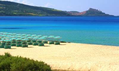 Spiaggia sabbiosa con mare cristallino, ombrelloni chiari e colline in lontananza.