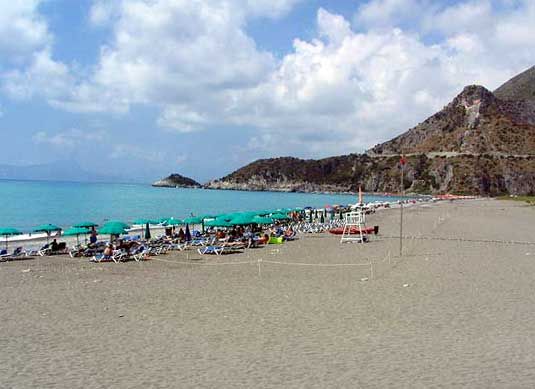 Spiaggia con lettini e ombrelloni, mare azzurro e colline sullo sfondo.