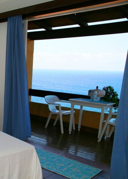 Interno con tende blu, tavolino e sedie su un balcone affacciato sul mare.