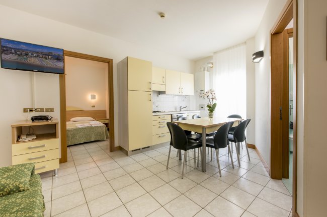 Soggiorno moderno con cucina beige, tavolo e sedie nere. TV appesa e accesso alla camera da letto.