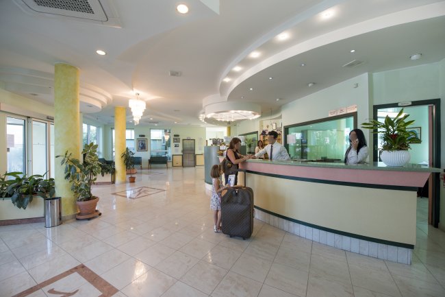 Reception di un hotel con persone al banco e piante decorative intorno.