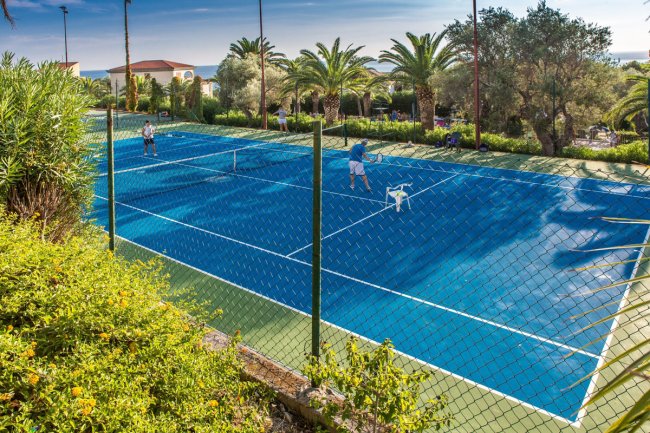 Campo da tennis blu con giocatori, circondato da palme e arbusti.