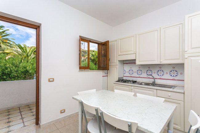 Una cucina con tavolo bianco, sedie e portafinestra aperta su un giardino.