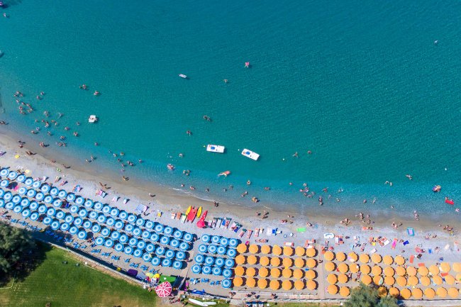 Spiaggia affollata vista dall'alto, con ombrelloni blu e arancioni lungo la costa sabbiosa.