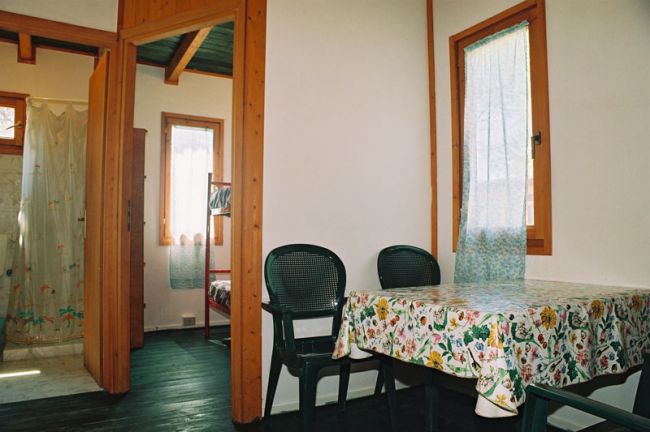 Interno di una casetta in legno con tavolo, sedie verdi, e vista su una camera con letto a castello.