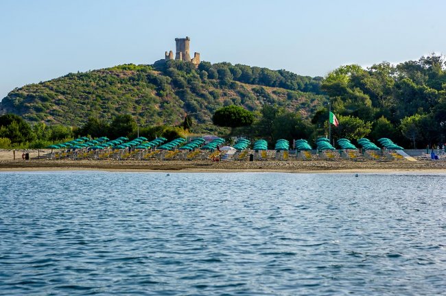 Spiaggia con file ordinate di ombrelloni verdi e una torre su una collina sullo sfondo.