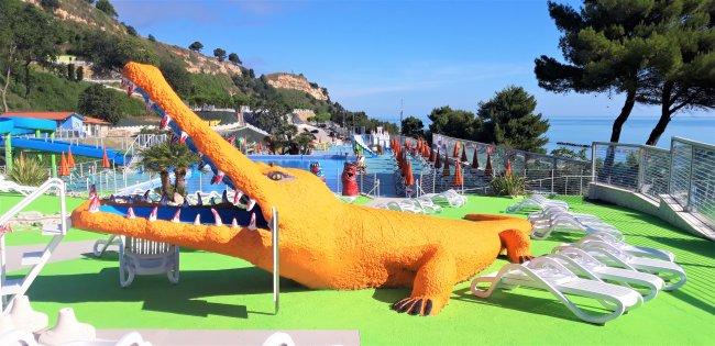 Grande statua di coccodrillo arancione accanto a lettini prendisole e ombrelloni.