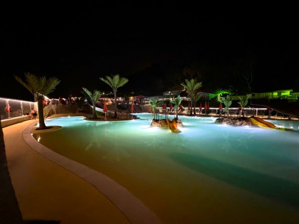 Piscina con illuminazione notturna e scivoli, palme sparse attorno.