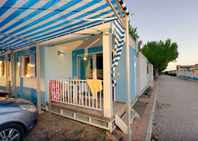Esterno di un bungalow azzurro con veranda e auto parcheggiata; un vialetto di ghiaia a lato.