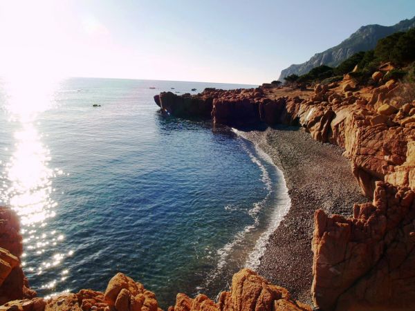 Spiaggia di ciottoli con scogliere rosse, mare blu e sole che riflette sull'acqua.