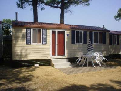 Casa mobile beige con tende blu, porta rossa, patio con tavolo bianco e sedie, sotto alberi.