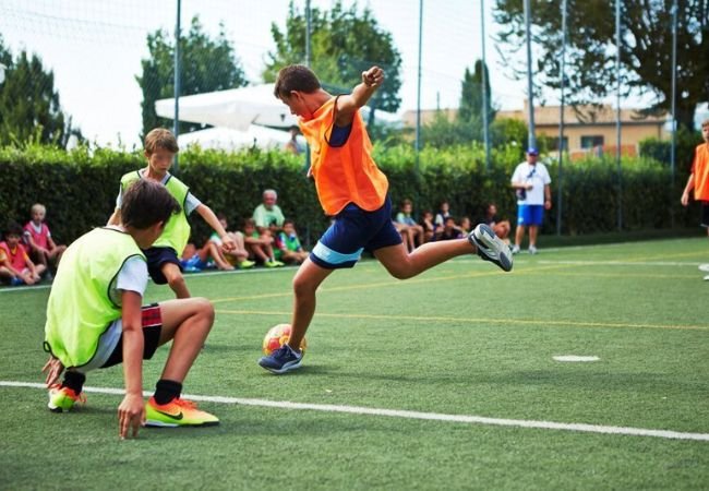 Ragazzi giocano a calcio su un campo in erba sintetica con maglie sportive colorate.
