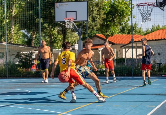 Giovani giocano a basket su un campo all'aperto, alcuni a torso nudo, con un canestro sullo sfondo.